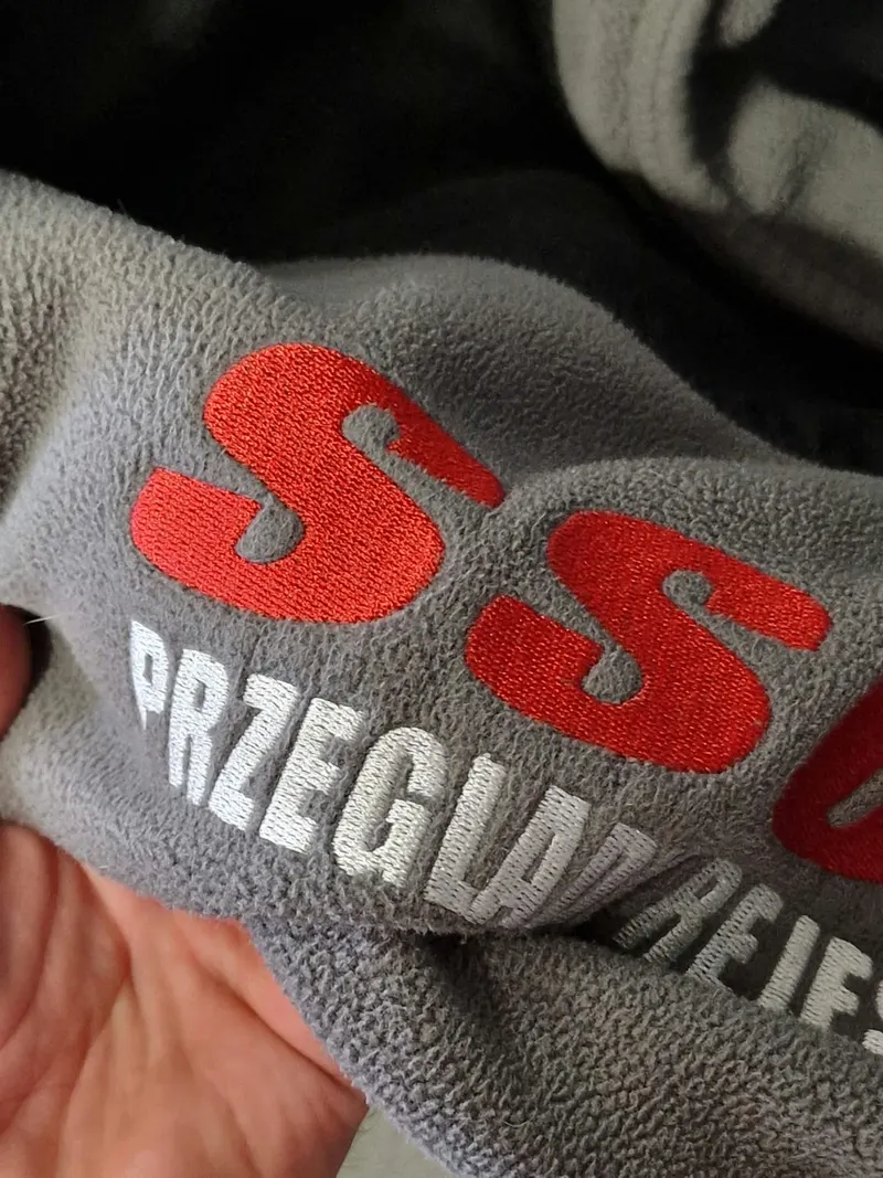 Zbliżenie haftowanego logo na szarym polarze po czterech latach codziennego noszenia w pracy