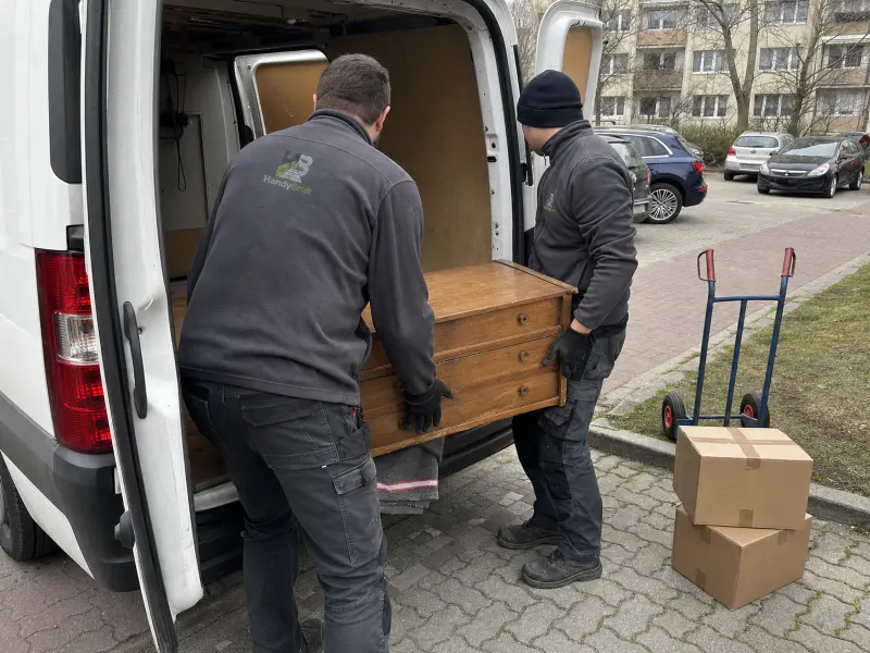 Dwóch pracowników w odzieży z logo HandyBruk przy załadunku komody do busa